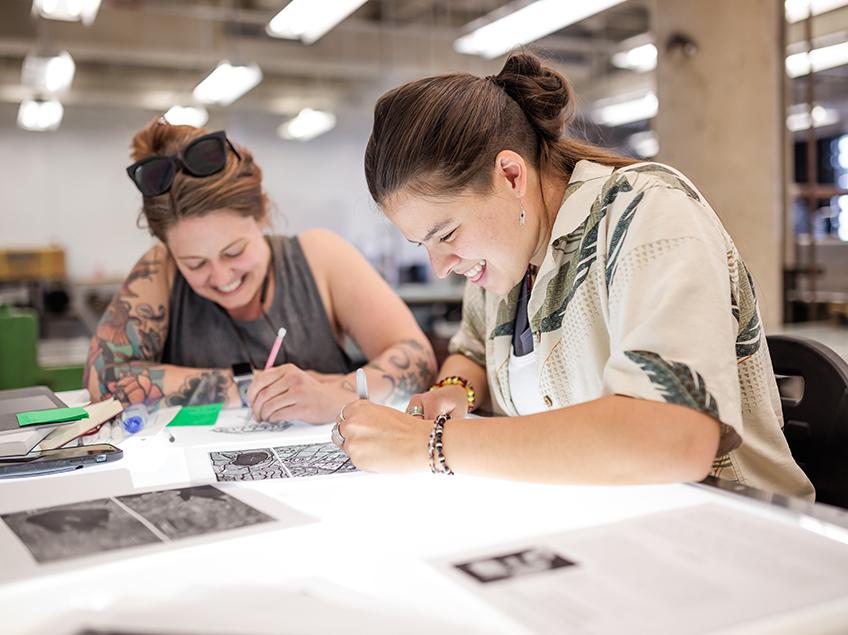 Two students smiling and laughing while working on the light table in the Print Studio.