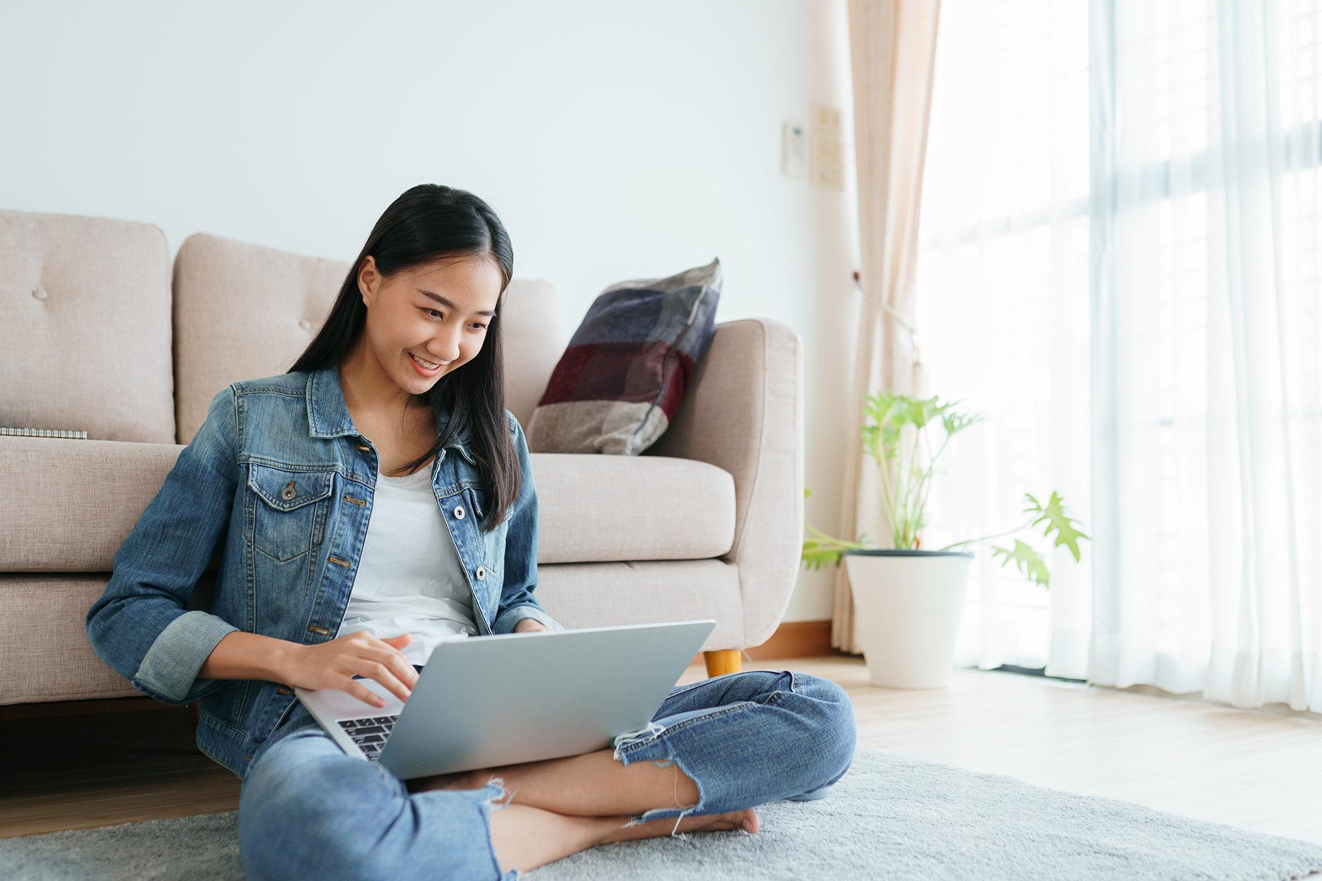 Asian girl wearing jeans using a laptop while sitting on the floor at home. Freelance girls are video conferencing with colleagues on social media. 概念工作在家和新常态